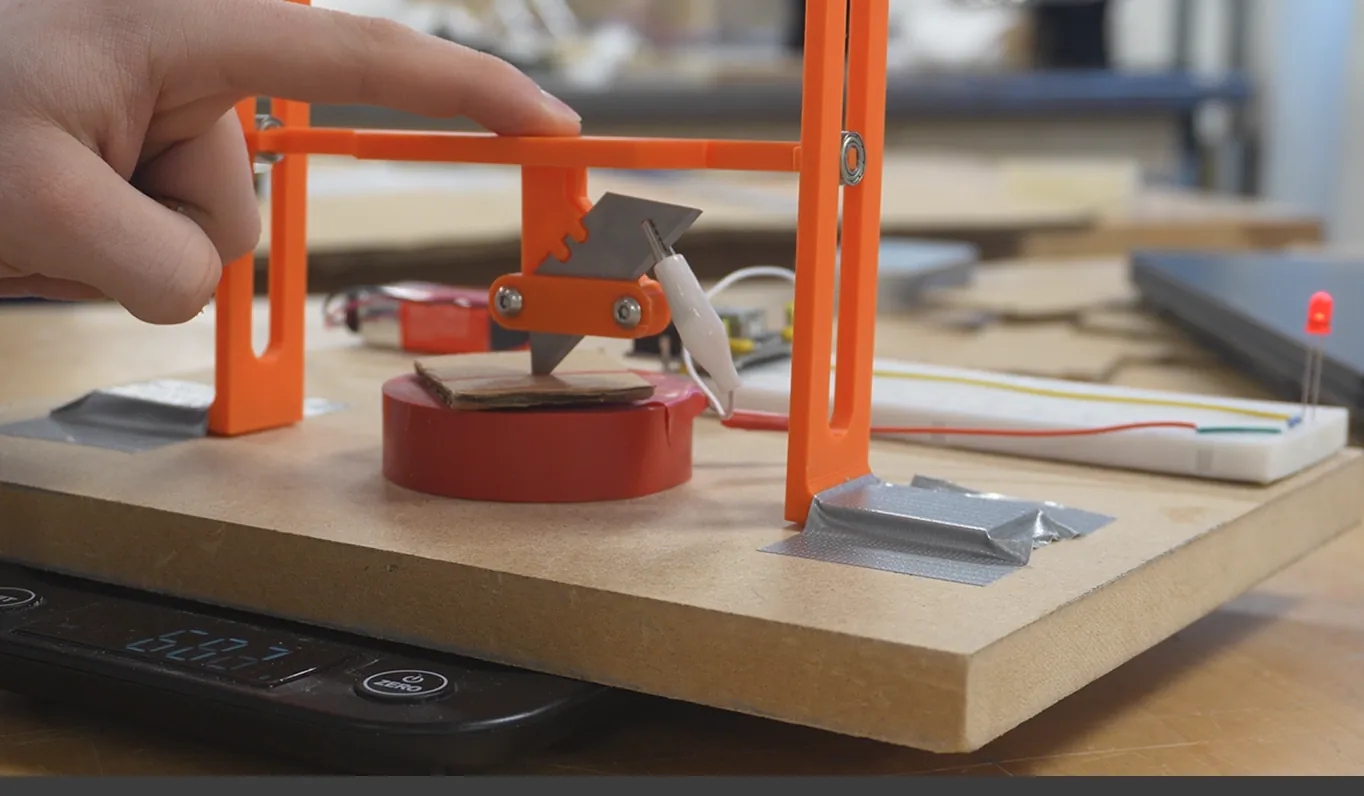 Piercing force rig: a blade on an orange 3D-printed frame pressed into cardboard on a digital scale, with an LED circuit that lights on contact