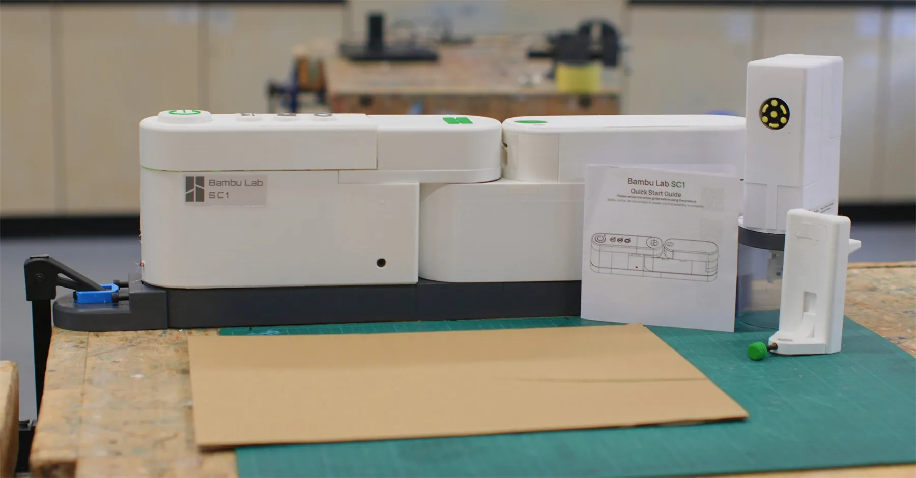 The SwivelCut prototype clamped to a workshop bench, with the cutting and tracing heads, a quick start guide and a sheet of cardboard on a cutting mat